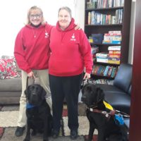 Therapy Dog Clive and Guide Dog Nate are sitting in front of Cheryl and Nancy who are wearing red ACB hoodies.